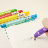 Stylo gomme violet en train d’effacer un trait de crayon sur un carnet, avec d’autres stylos gomme en arrière-plan