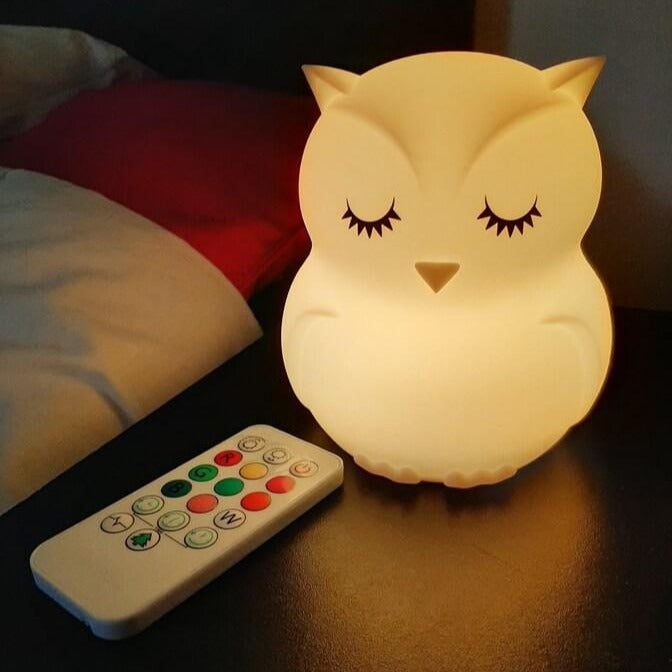 Veilleuse hibou à LED allumée en blanc sur une table de chevet, avec sa télécommande